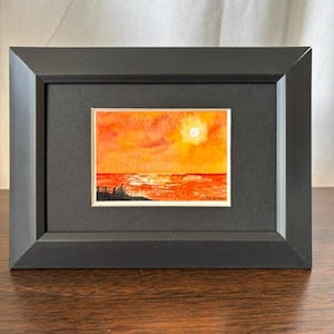 May include: A small rectangular watercolor painting of a sunset over the ocean, framed in a black frame with a black mat. The painting features vibrant orange and yellow hues, a bright sun, and a dark silhouette of a fence along the shoreline.