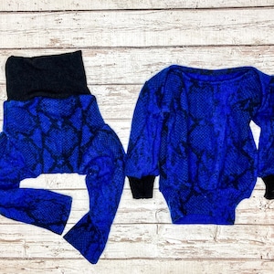 May include: A baby outfit set featuring a blue snakeskin print. The set includes a long-sleeved bodysuit with black cuffs and a pair of pants with a black waistband. The outfit is made of a soft, textured fabric.