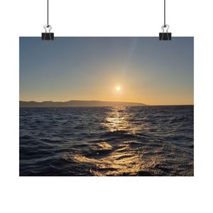 Può includere: Una fotografia paesaggistica di un tramonto sull'oceano. Il sole è un luminoso globo dorato basso nel cielo, che proietta un riflesso scintillante sull'acqua blu scuro increspata. Una sagoma scura di una costa è visibile all'orizzonte.