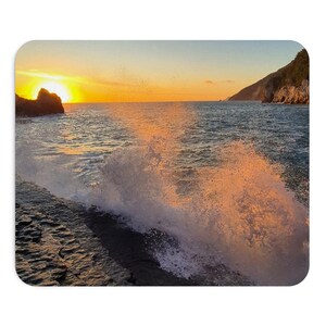 Può includere: Un tappetino per mouse rettangolare con una scena costiera al tramonto. L'immagine mostra rocce scure e strutturate in primo piano con le onde dell'oceano che si infrangono contro di esse, illuminate da un sole arancione dietro una scogliera.