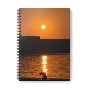 May include: A spiral-bound notebook featuring a cover image of a bright orange sunset over a body of water. The sun is positioned high in the sky, casting a shimmering golden reflection across the rippling water surface toward a dark silhouette.