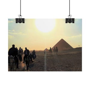 Può includere: Una fotografia paesaggistica che mostra le sagome di persone a cavallo di cammelli su un terreno desertico sabbioso. Sullo sfondo, le piramidi di Giza sono visibili sotto un sole luminoso. La scena cattura un'escursione nel deserto al tramonto.