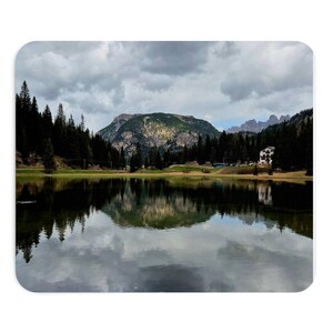 Può includere: Una vista paesaggistica di un lago calmo che riflette una grande montagna e una fitta foresta di alberi sempreverdi sotto un cielo nuvoloso e coperto. Un edificio bianco con dettagli scuri è visibile sul lato destro vicino al bosco.