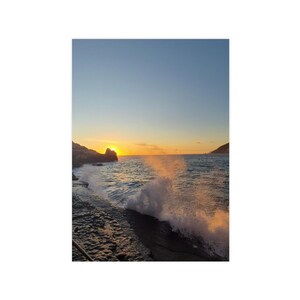 Può includere: Una scena costiera al tramonto con rocce scure e frastagliate in primo piano e onde dell'oceano che si infrangono contro di esse. Il sole dorato è basso all'orizzonte, proietta una luce calda sull'acqua e crea spruzzi di schiuma marina bianca.