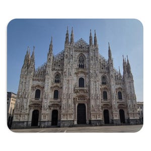Può includere: Il Duomo di Milano è una grande cattedrale in marmo bianco caratterizzata da una complessa architettura gotica, numerose guglie appuntite, dettagliate sculture in pietra e molteplici ingressi ad arco sotto un cielo azzurro.