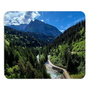 Può includere: Una vista paesaggistica di una catena montuosa sotto un cielo blu con nuvole bianche. Un fiume scorre attraverso una valle piena di fitti boschi di pini. Una stretta strada sterrata costeggia il fiume verso la base delle montagne.