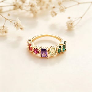 May include: A gold-toned ring featuring a row of multi-coloured gemstones in various shapes, including emerald, oval, and rectangular cuts. The stones display a vibrant spectrum of colours, including red, purple, yellow, and green, set in a polished metal band.