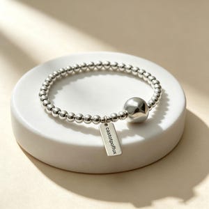 May include: A silver-toned beaded bracelet featuring a series of small, uniform spherical beads, one larger smooth round bead, and a rectangular metal charm dangling from the band. The charm is engraved with the text castotpoffux in black lettering.