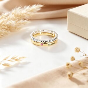 May include: A two-tone stacked ring set featuring a silver band engraved with the Roman numerals VII.XXIV.MMXXI and a small blue stone, paired with a gold band engraved with the name Emma and a small pink stone set into the metal surface.
