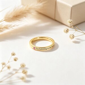 May include: A gold-toned band ring featuring a small, round, pink gemstone set into the metal. The outer surface of the ring is engraved with the black text "I'm listening to my heart" positioned next to the pink stone on the smooth, polished band.