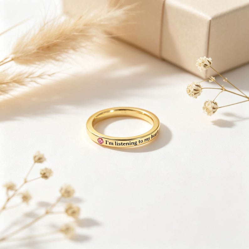 May include: A gold-toned band ring featuring a small, round, pink gemstone set into the metal. The outer surface of the ring is engraved with the black text "I'm listening to my heart" positioned next to the pink stone on the smooth, polished band.