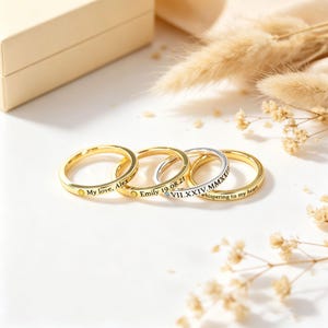 May include: Four thin metal rings arranged in a row. Three rings are gold-toned and one is silver-toned. Each ring features custom engraving and a small embedded gemstone. The engravings include the text My love, Alex, Emily 19.08.21, and VII.XXIV.MMXXIV, whispering to my heart.