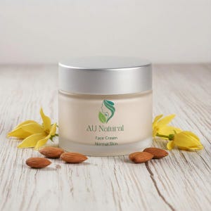 Peut inclure: Un pot en verre rond de crème pour le visage avec un couvercle argenté. L'étiquette présente un logo avec une feuille verte et un profil, avec le texte AU Natural, Face Cream et Normal Skin. Le pot est entouré de fleurs jaunes et d'amandes.