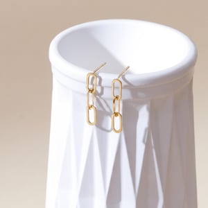 May include: A pair of gold-toned drop earrings featuring a delicate chain design with three elongated oval links. The earrings have a post-back closure and a polished metallic finish, suspended from the rim of a white cylindrical object.