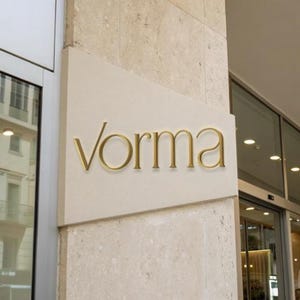 Puede incluir: Un letrero rectangular de piedra beige montado en el exterior de un edificio con la palabra "vorma" en letras metálicas doradas en relieve de estilo serif. El letrero está colocado entre dos ventanas de cristal con marcos de metal.