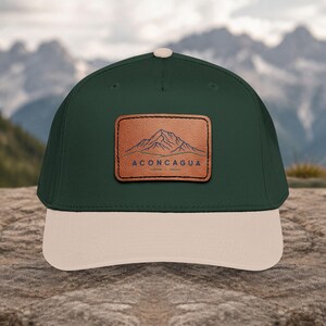 May include: A dark green baseball cap with a light beige brim and a top button. A rectangular tan leather patch is centered on the front, featuring a blue line drawing of a mountain range above the text ACONCAGUA and a small decorative line.