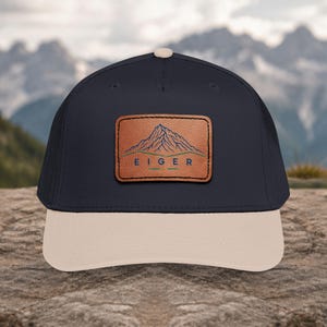 May include: A navy blue baseball cap with a beige brim and a top button. Centered on the front is a rectangular brown leather patch featuring a blue line drawing of a mountain peak above the text EIGER in blue, with green horizontal lines.