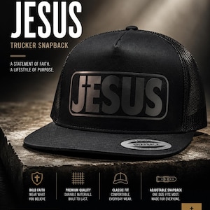 May include: A black trucker snapback hat featuring the word JESUS in a bold, dark grey font on the front panel. The hat has a mesh back, a flat brim, and a circular sticker on the brim that reads THE AUTHENTIC SNAPBACK YUPOONG SINCE 1974.