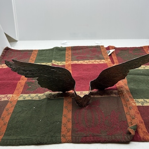 Op de afbeelding: Een metalen, donkerkleurig decoratief object in de vorm van uitgestrekte vleugels. De vleugels zijn voorzien van gedetailleerde, geëtste veerpatronen en zijn verbonden bij een centrale, ronde basis met een uitgesneden ontwerp en verweerde afwerking.