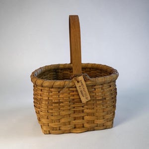 May include: A woven brown basket with a single upright wooden handle. The basket features a rectangular base and rounded corners. A small, rectangular tan tag is tied to the handle with twine, displaying the text HANDCRAFTED BY R J SHARP.