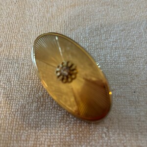 May include: An oval-shaped gold-tone brooch featuring a sunburst pattern radiating from the center. A small, circular, flower-shaped metal embellishment with a clear, faceted stone is positioned directly in the middle of the brooch surface.