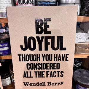 May include: A rectangular tan sign featuring bold black text that reads BE JOYFUL THOUGH YOU HAVE CONSIDERED ALL THE FACTS, followed by the name Wendell Berry and a small black graphic illustration of a person resting in a hammock.