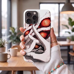 May include: A smartphone case featuring a graphic illustration of a pale hand forming a circle with fingers, revealing a sharp-toothed mouth inside the circle. The background displays multiple glowing red, swirling eyes against a white textured surface.