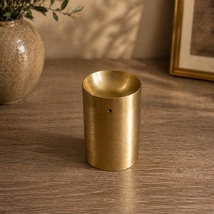 Peut inclure: Un porte-encens cylindrique en laiton avec une finition métallique brossée. L'objet présente une large ouverture supérieure concave et un petit trou circulaire unique positionné sur la partie supérieure de la paroi extérieure pour l'encens.