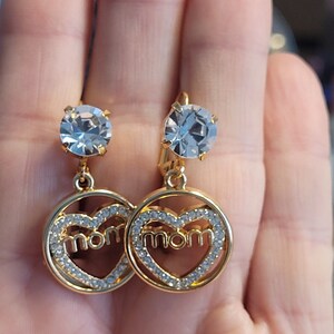 Può includere: Un paio di orecchini color oro con una gemma rotonda e trasparente nella parte superiore. Ogni orecchino presenta un ciondolo a forma di cuore con la parola "mamma" al centro, delineato da piccoli cristalli trasparenti. Chiusura a leva.