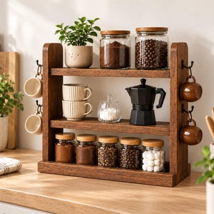 May include: A three-tiered wooden kitchen organiser featuring two side panels with four black metal hooks for hanging mugs. The shelves hold glass jars with wooden lids, two ribbed ceramic mugs, a glass sugar bowl, and a black moka pot.