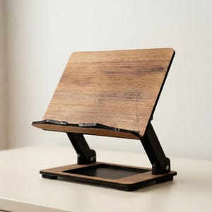 May include: A foldable tablet or book stand constructed from wood with a visible grain pattern. The stand features a rectangular support panel, a flat base, and black metal hinges that allow for adjustable positioning of the viewing angle.