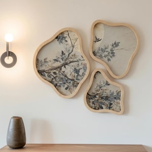 May include: Three irregular, organic-shaped wall art pieces with light wood frames. Each piece features a printed illustration of tree branches with leaves and small birds in muted tones of gray, brown, and cream, arranged in a cluster on the wall.