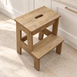 May include: A two-step wooden stool with a light brown oak wood grain finish. The stool features a rectangular top step with a central oval cutout handle and a lower step. The structure is held together by visible metal screws at each corner.