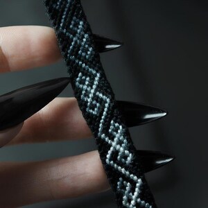 May include: A woven friendship bracelet featuring a repeating geometric pattern in light blue thread against a solid black background. The band is constructed from tightly knotted embroidery floss, creating a textured surface with a consistent width.