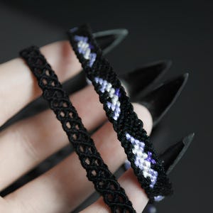 May include: Two handmade bracelets made of black thread. One bracelet features a delicate, repeating open-work lattice pattern. The second bracelet is crafted with a solid black base and a diagonal, woven geometric design in shades of purple and white.