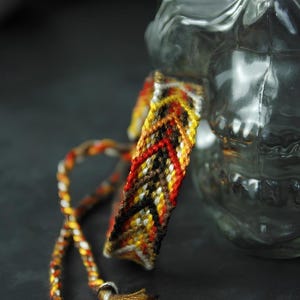 Puede incluir: Una pulsera de la amistad tejida con un patrón de chevrón en tonos rojo, naranja, amarillo, marrón, negro y blanco. La pulsera incluye largos lazos trenzados que terminan en pequeñas borlas de hilo multicolor en cada uno de los dos extremos.