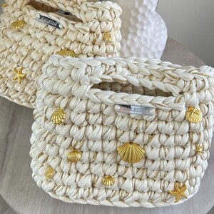 May include: Two cream-colored crocheted handbags featuring integrated handles and gold-toned metallic shell and starfish charms. Each bag displays a silver-toned rectangular label with the text KALEIDA attached to the front upper section.