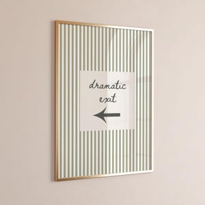 May include: A rectangular wall art piece in a thin gold frame featuring a vertical sage green and white striped background. Centered is a white square containing the black handwritten text "dramatic exit" above a black left-pointing arrow.
