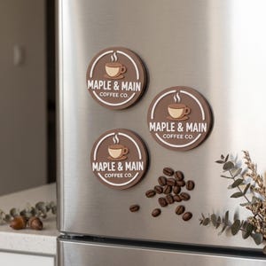 Peut inclure: Trois aimants circulaires marron et blancs arborant le dessin d'une tasse de café fumante et le texte Maple & Main Coffee Co. Les aimants sont disposés sur une surface métallique à côté d'un petit groupe de grains de café torréfiés foncés.
