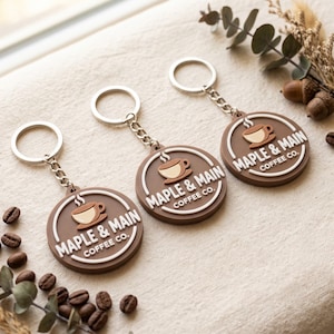 May include: Three circular brown keychains featuring a white raised logo for Maple & Main Coffee Co. Each keychain includes a silver metal ring and chain, with a graphic of a steaming coffee cup centered above the white text lettering.