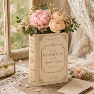 May include: A rectangular beige book-shaped vase featuring decorative gold-toned borders and the text "OUR FOREVER STORY," "Written By Amelia & James," "EST. 09.21.2027," and "LOVE WRITES THE BEST STORIES." It holds a bouquet of pink peonies and ranunculus.