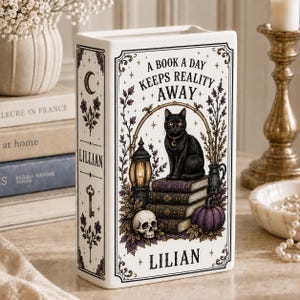 May include: A white ceramic book-shaped vase featuring a black cat sitting on a stack of books, a skull, a pumpkin, and a lantern. The text reads A BOOK A DAY KEEPS REALITY AWAY and LILIAN. The design includes purple floral accents and a crescent moon.