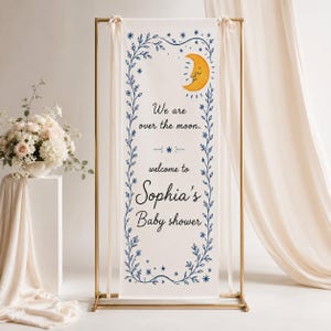 May include: A vertical white fabric banner suspended on a gold metal frame. The banner features a blue vine border, a yellow crescent moon with a face, and the text: We are over the moon. welcome to Sophia's Baby shower. Blue stars accent the design.