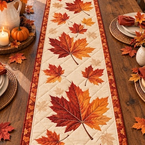 May include: A rectangular quilted table runner featuring a cream-colored center with a repeating pattern of vibrant orange, red, and yellow maple leaves. The runner is bordered by a dark orange fabric decorated with a smaller maple leaf print.