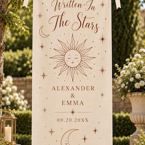 May include: A rectangular off-white fabric banner hanging from a rope with cream-colored ribbons. It features brown printed celestial graphics including a sun, a crescent moon, and stars, with the text Written In The Stars, ALEXANDER & EMMA, 09.20.20XX.