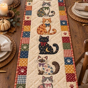 May include: A quilted table runner featuring a cream-colored center panel with six illustrated cats in various colors and floral patterns, each wearing a bow. The border consists of a patchwork of small squares in red, blue, yellow, and green.