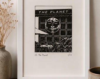 Puede incluir: Una impresión en linóleo en blanco y negro dentro de un marco blanco que muestra la fachada de un café con el texto THE PLANET y un icono de una luna con rostro sobre una mesa y sillas. Incluye el texto 03. The Planet y el número 1/15.