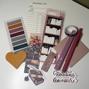 May include: A collection of stationery items including a reading list notepad, a bookshelf bookmark, a floral bookmark, a heart-shaped yellow leather corner bookmark, a roll of patterned washi tape, a mauve highlighter, a pen, and two stickers.