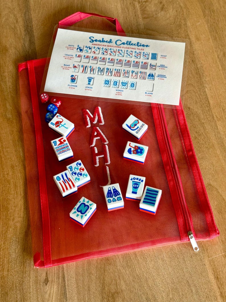 Pu&ograve; includere: Una custodia rossa in rete con cerniera contenente tessere e dadi Mahjong. Le tessere sono bianche con grafica e testo colorati, tra cui "Joker" e "SPF". Un foglio di plastica trasparente mostra i tipi di tessere. La custodia ha un manico rosa e la parola "MAH" ricamata.
