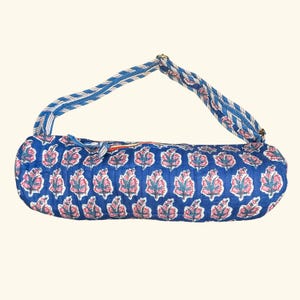 May include: A cylindrical quilted yoga mat bag in a deep blue color featuring a repeating floral pattern in pink and white. The bag includes a long, adjustable shoulder strap with a blue and white striped design and a zipper closure along the top.